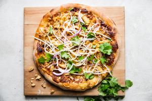 Thai style chicken pizza with cheese. onions, hoisin sauce peanuts and cilantro on a cutting board