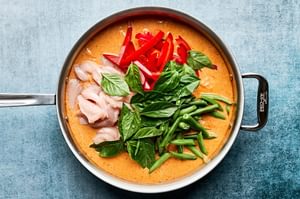 a pan of creamy thai red curry with chicken and vegetables being added