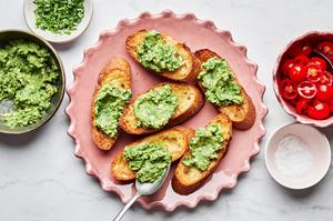 toasted baguette topped with mashed avocado surrounded by a bowl of tomato bruschetta, flaky salt and chopped basil