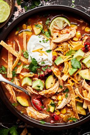a bowl of chicken tortilla soup topped with tortilla strips, cilantro, sour cream, cheese, avocado and lime juice