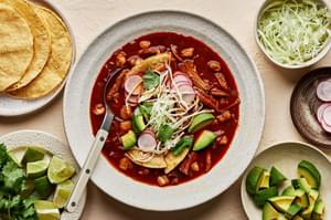 a bowl of homemade turkey pozole rojo topped with seasoned cabbage, avocado, cilantro, radishes, and lime juice.