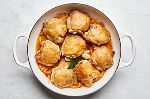 Browned bone-in, skin-on, chicken cooking in vinegar with oranges, bay leaf and olives in a white braiser.