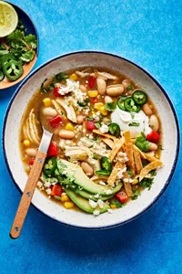 a bowl of white bean chicken chili topped with jalapeño, Sour cream, avocado, tortilla strips, cotija and cilantro