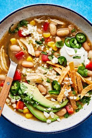 a bowl of white bean chicken chili topped with jalapeño, Sour cream, avocado, tortilla strips, cotija and cilantro