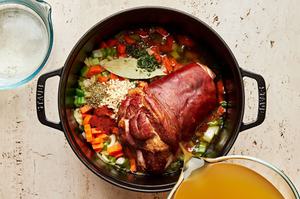 water and chicken broth being poured in a pot with onion, celery, carrots, spices, and ham hock