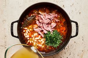 chicken broth and parsley being added to a pot of white bean and ham soup