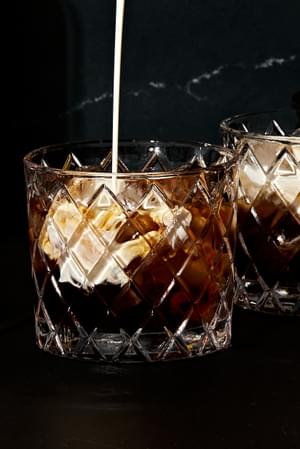 heavy cream being poured into a cocktail glass with vodka, coffee liqueur and ice