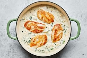 chicken being added to a skillet with spinach artichoke cream sauce